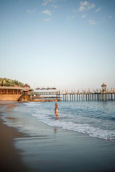 A woman in a swimsuit and a hat with glasses walks along the beach at sunset. The concept of sea recreation. Selective focus