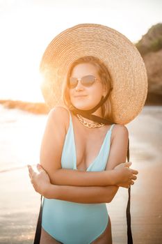 A young tanned woman in a beautiful swimsuit with a straw hat stands and rests on a tropical beach with sand and looks at the sunset and the sea. Selective focus. Vacation concept by the sea
