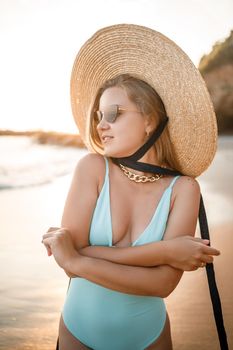 A young tanned woman in a beautiful swimsuit with a straw hat stands and rests on a tropical beach with sand and looks at the sunset and the sea. Selective focus. Vacation concept by the sea
