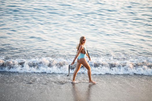 Beautiful girl in a blue swimsuit and hat on a sandy beach at the sea in the sunset sunlight