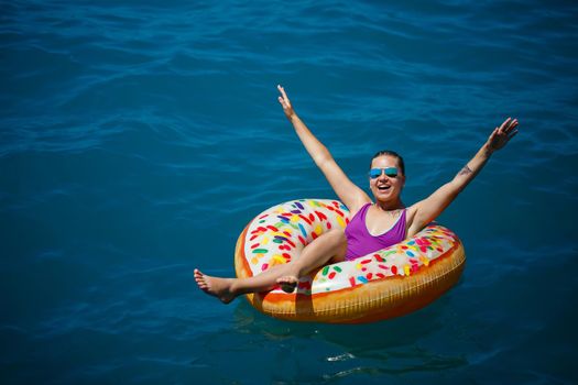 A young beautiful girl in a bright swimsuit lies on a large inflatable ring and floats on the blue sea on a bright sunny summer day