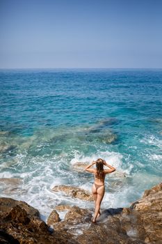 Sexy woman in full body swimsuit with long hair walks on large rocks on a rocky beach during a storm at sea. Back view. Woman swimwear island tropics landscape exotic walk