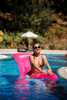 Sexy woman in sunglasses with a smile on her face in a swimsuit lies on a pink inflatable mattress in the pool. Relax by the pool on a hot summer sunny day. Vacation concept