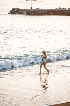 Beautiful girl in a blue swimsuit and hat on a sandy beach at the sea in the sunset sunlight