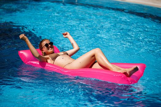 Sexy woman in a swimsuit lies on a pink inflatable mattress in the pool. Relax by the pool on a hot summer sunny day. Vacation concept