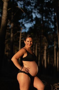 A pregnant woman caresses her belly and smiles at camera wearing black underwear in the nature during sunset