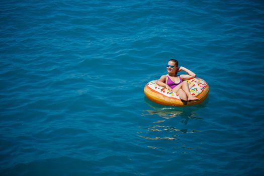 A young beautiful girl in a bright swimsuit lies on a large inflatable ring and floats on the blue sea on a bright sunny summer day