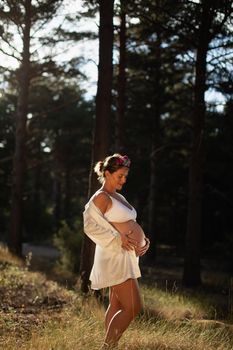 A pregnant woman caresses her belly and her future child wearing a silk robe in the park during sunset