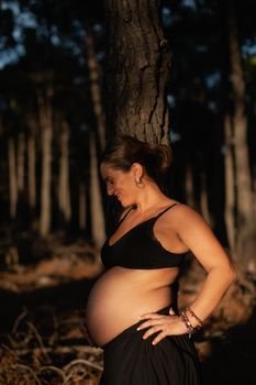 A pregnant woman caresses her belly and smiles wearing black underwear in the nature during sunset