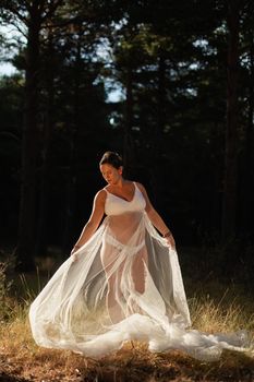A pregnant woman poses holding part of her wedding dress with her hands in the nature amongst beautiful sunset lights