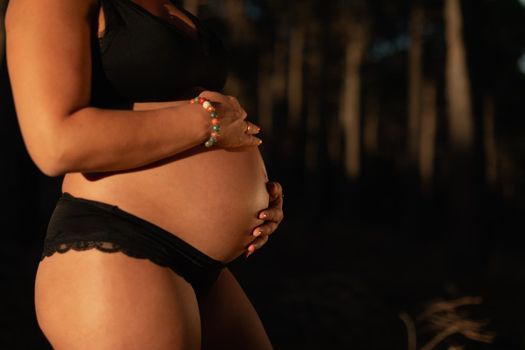 Close-up portrait of a pregnant woman caressing her belly wearing underwear in the park during sunset