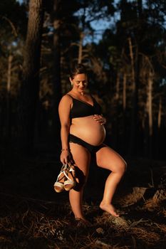 A pregnant woman caresses her belly and smiles while holing her shoes in the park during a beautiful sunset