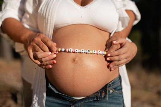 Close-up of the hands of a young couple, holding a bead chain over mom's belly with the name of their future child