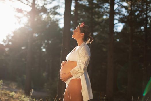 A pregnant woman caresses her belly and her future child wearing a silk robe in the park during sunset