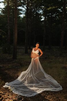 A pregnant woman poses caressing her belly wearing part of her wedding dress in the nature during sunset