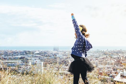 side view of a little child girl posing powerful with her fist in the air disguised as a superhero with homemade costume in front of a cityscape, imagination and girl power concept