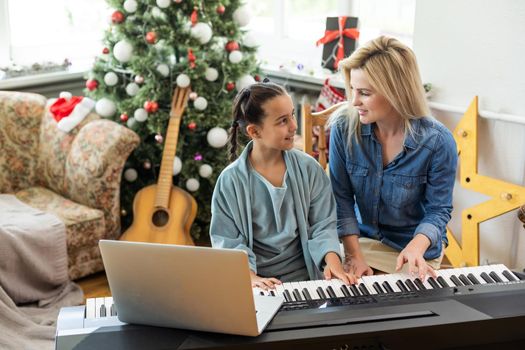 Portrait of a little girl playing the piano decorated with Christmas ornaments. Happy mother smiling on the background. Winter and New Year holidays.