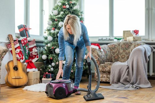 Young woman cleaning with vacuum cleaner, vacuuming under Christmas Tree needles with New Years ornaments on hardwood wooden floor