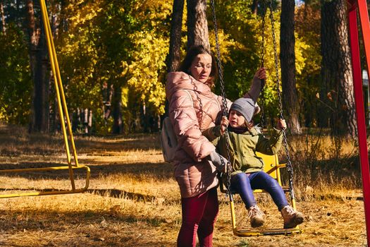 a woman in relation to her child or children. Young cute mother shakes her baby daughter on a swing in a bright autumn park.