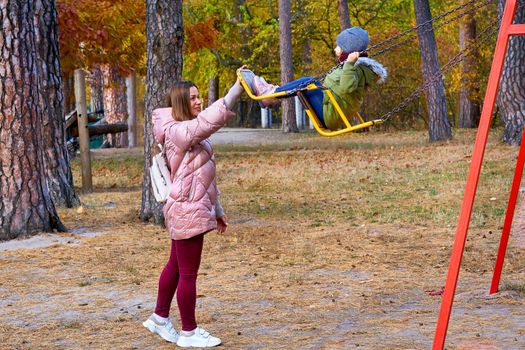 a woman in relation to her child or children. Young cute mother shakes her baby daughter on a swing in a bright autumn park.