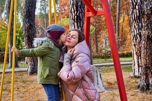 a woman in relation to her child or children. Cute child gently kisses and hugs her mom on a swing in the autumn forest park