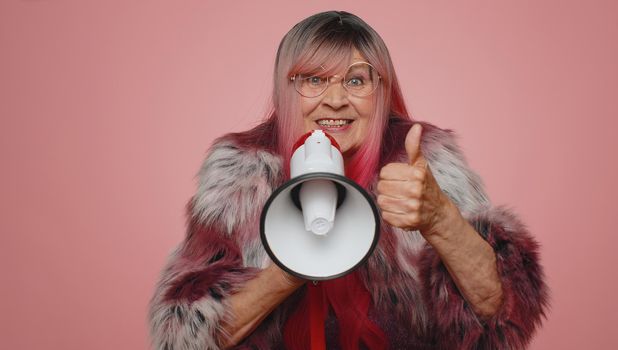 Smiling happy stylish old woman talking with megaphone, proclaiming news, loudly announcing sale advertisement, warning using loudspeaker to shout speech. Senior elderly grandmother on pink background