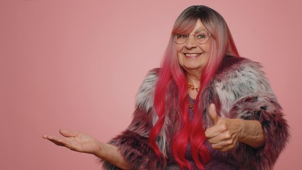 Lovely stylish old woman showing thumbs up and pointing empty place, advertising area for commercial text, copy space for goods promotion. Senior elderly grandmother. Studio indoors on pink background