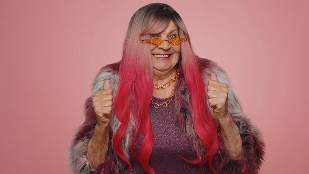 Happy joyful old woman in coat shouting, raising fists in gesture I did it, celebrating success, winning, birthday, lottery goal achievemen. Elderly grandmother indoors on pink studio wall background