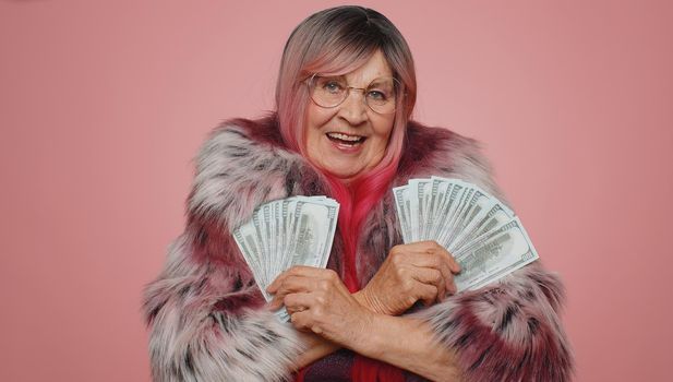 Happy stylish woman holding fan of cash money dollar banknotes celebrate dance, success business career, lottery game winner, big income, wealth. Senior elderly grandmother isolated on pink background