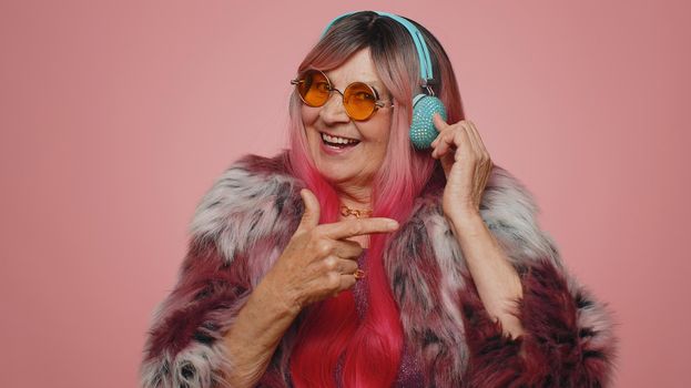 Funny old woman 70s listening music, smiling dancing to disco party music rhythmically moving his hands, having fun. Senior grandmother celebrating winning, birthday isolated on pink wall background