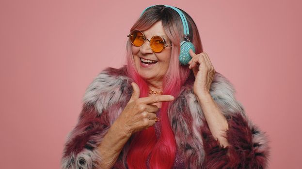 Funny old woman 70s listening music, smiling dancing to disco party music rhythmically moving his hands, having fun. Senior grandmother celebrating winning, birthday isolated on pink wall background