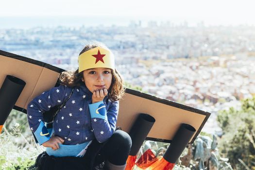 funny little child girl poses disguised as a superhero with homemade costume and cardboard plane wings in front of a cityscape, imagination and girl power concept