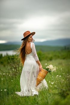 A middle-aged woman in a white dress and brown hat stands on a green field and holds a basket in her hands with a large bouquet of daisies. In the background there are mountains and a lake