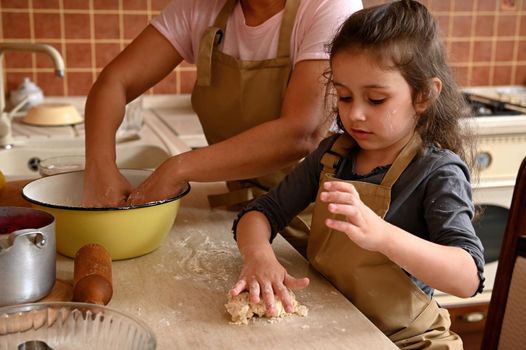 Adorable mischievous European little girl, lovely daughter in beige chef apron, kneading dough on a kitchen table, learning cooking homemade pastries, cherry pie and festive tartlets with her mom.