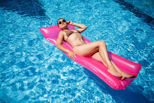 Sexy woman in a swimsuit lies on a pink inflatable mattress in the pool. Relax by the pool on a hot summer sunny day. Vacation concept