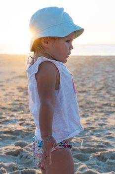 Little blonde girl in a panama hat stands with her back on the beach and looks at the ocean