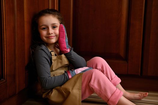 Portrait of an adorable Caucasian child girl, little baker confectioner in a beige chef apron and kitchen mittens, sitting on the floor and leaning against wooden kitchen cupboard, smiling to camera