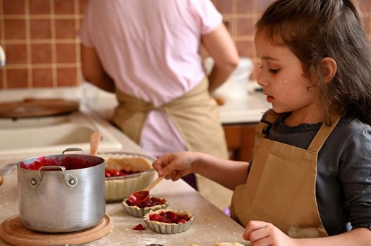 Adorable little girl in chef's apron, filling molds with rolled dough and cherries, preparing tartlets according to family recipe. Handsome playful kid learns cooking while helping her mom in kitchen