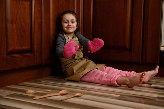 Charming European little girl, adorable child baker confectioner wearing beige chef's apron, handing her hands in kitchen mittens to camera while sitting barefoot on the floor and smiling a cute smile