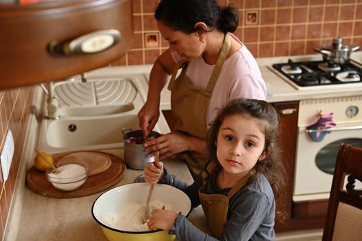 Beautiful Caucasian preschool child, adorable little girl in a chef's apron, daughter helps to knead the dough while her mom prepares caramelized cherries to fill tartlets, enjoying pastime together