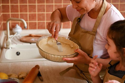Close-up. Selective focus on Hispanic stunning housewife using a fork, piercing the rolled out dough in a baking mold while preparing a festive homemade pie. Mom and daughter cook tartlets together