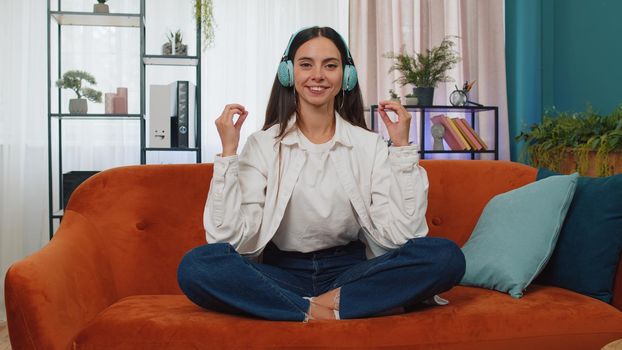 Keep calm down, relax, inner balance. Adult girl breathes deeply with mudra gesture, eyes closed meditating with concentrated thoughts, peaceful mind. Young woman sit at home in living room on couch