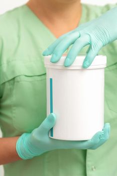 Hands in protective gloves of beautician open a white body cream jar on white background