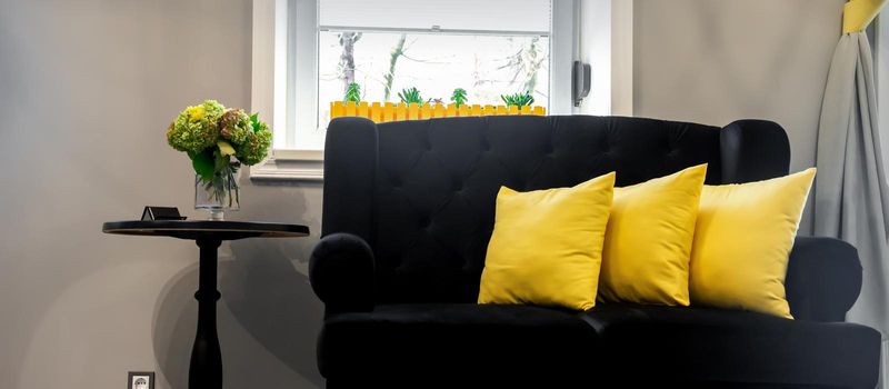Interior of the beauty room. Black sofa with small yellow pillows and coffee table on a gray wall background with a window