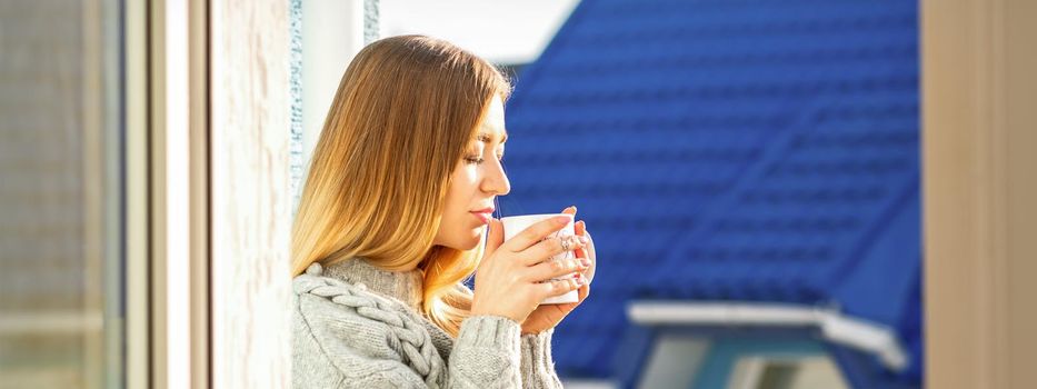 Woman relaxing on the balcony with coffee. A beautiful young woman in a sweater holds a cup of coffee standing near the window in the morning