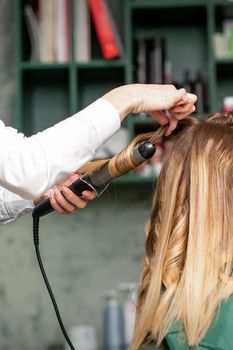 Creating curls with curling irons. Hairdresser makes a hairstyle for a young woman with long red hair in a beauty salon