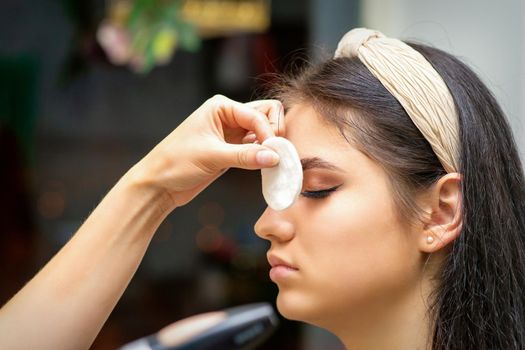 Side view of a makeup artist covers female eye with a cotton pad using airbrush making makeup foundation on her face in a beauty salon