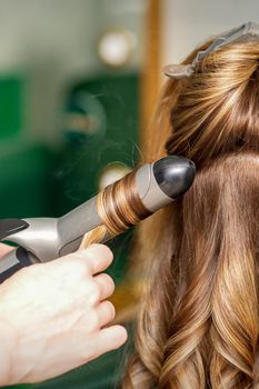 Creating curls with curling irons. Hairdresser makes a hairstyle for a young woman with long red hair in a beauty salon