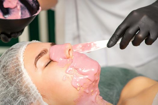 The cosmetologist applying an alginate mask to the face of a young woman in a beauty salon
