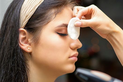 Side view of a makeup artist covers female eye with a cotton pad using airbrush making makeup foundation on her face in a beauty salon